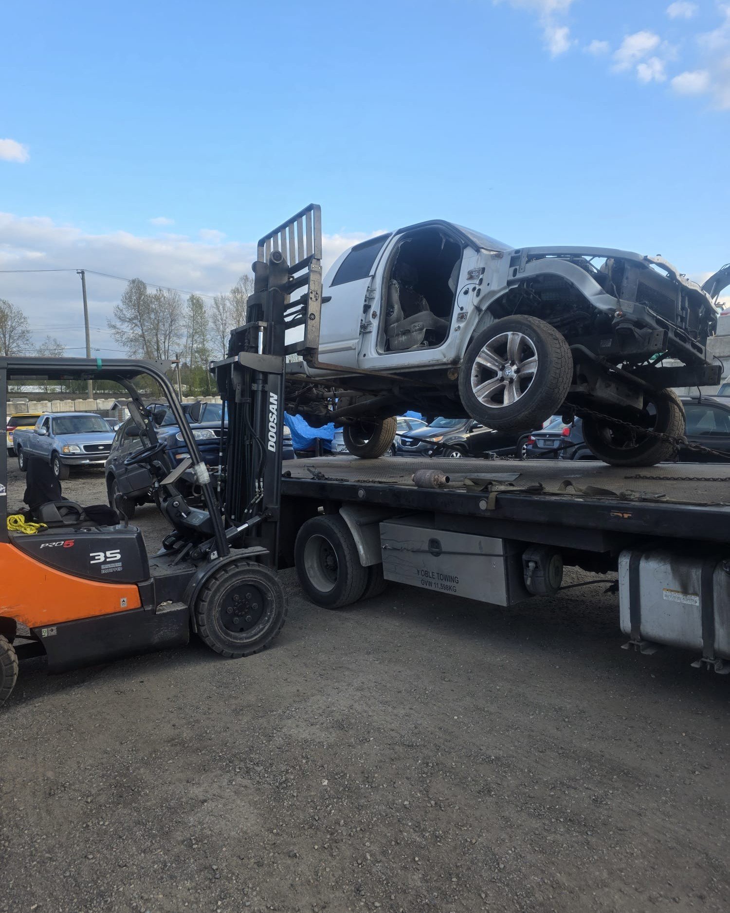 Dismantled white pickup secured on a flatbed trailer beside a forklift
